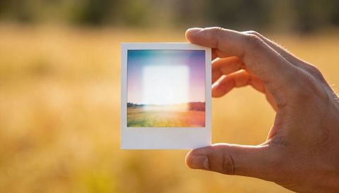 Holding polaroid frame over sunlit meadow during golden hour evoking vintage memory