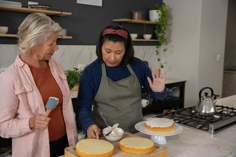 Senior women frosting layered cake together in cozy kitchen, baking friendship and teamwork