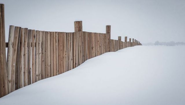 Weathered Wooden Fence Stretching Through Snowdrift on Winter Plain, Receding Posts