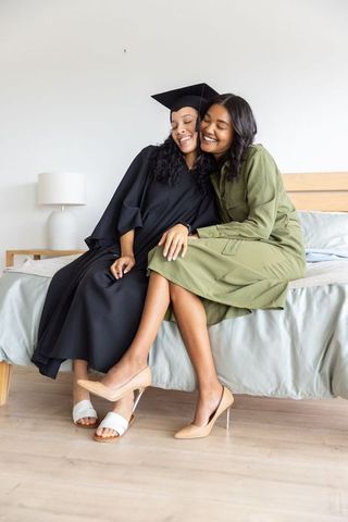 Diverse sisters celebrating graduation at home wearing gowns and caps on bed
