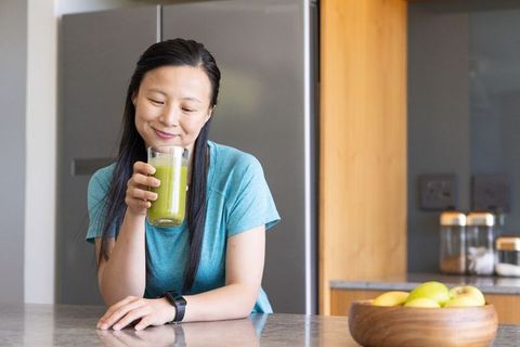 Smiling mature asian woman drinking green smoothie in modern kitchen wearing fitness watch