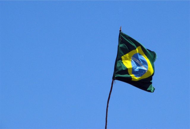 Brazilian flag waving high against clear blue sky, symbolizing national pride and patriotism. Ideal for use in educational materials, travel and tourism promotions, national celebrations, and cultural presentations.