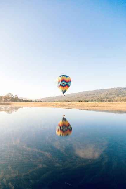 Hot air balloon floating over a calm lake with its colorful reflection visible on the water, under a clear blue sky. Mountains and nature in the background enhance the tranquility of the scene. Suitable for promoting travel destinations, adventure activities, peaceful nature escapes, or scenic photography.