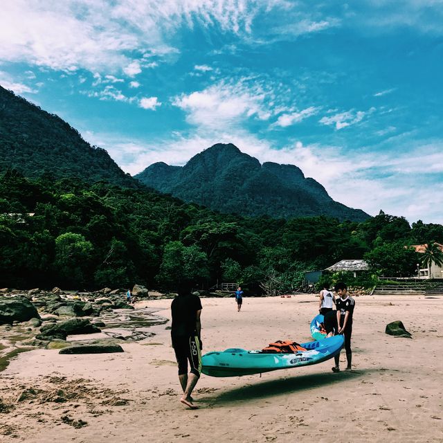 Several people are seen carrying a kayak along a sandy beach with lush green mountains in the background. The scene suggests outdoor activities and teamwork, ideal for promoting beachside adventures, outdoor gear, mountain retreats, or teamwork concepts.