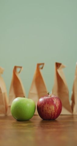 This depicts red and green apples sitting in front of a row of neatly packed paper lunch bags, set against a soft green background. Useful for themes related to healthy nutrition, apple as a snack, and meal prep or school lunches. Works well in articles or marketing that focus on diet tips, healthy living, educational content about nutrition, or promoting eco-friendly packaging solutions.