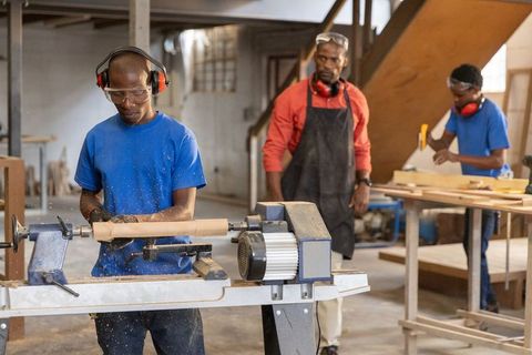 African american instructor mentoring teens using wood lathe and sanding plank in workshop