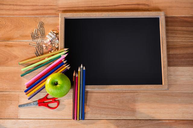 Chalkboard with colored pencils and green apple on wooden table. Ideal for educational materials, back-to-school promotions, classroom decor, and creative learning resources. Perfect for illustrating school-related themes, teaching aids, and art projects.