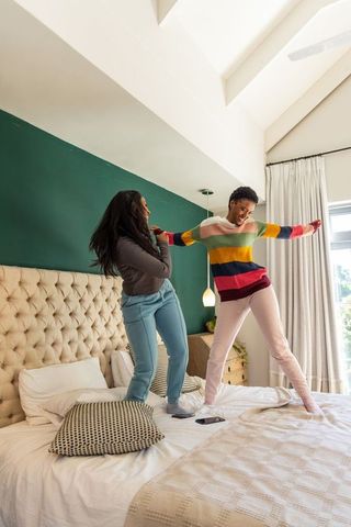 Playful diverse female friends jumping and balancing on king-size bed in bright bedroom