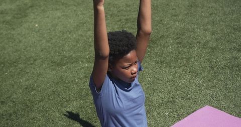 Young girl stretching arms overhead on field beside purple yoga mat sunny outdoor exercise