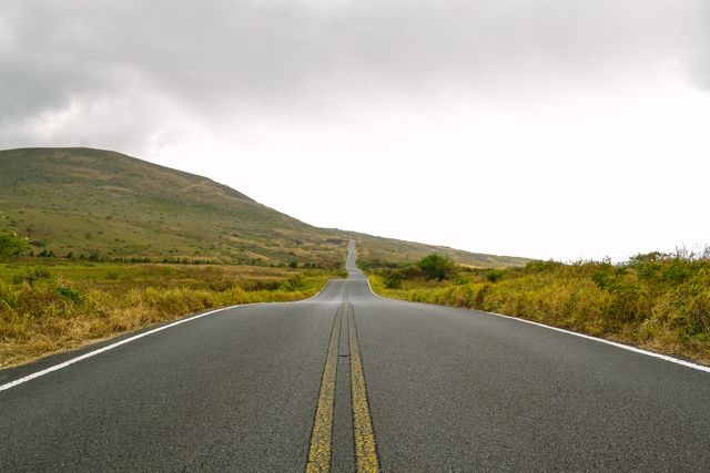 Photo of a long, empty road stretching towards distant hills under a cloudy sky. Asphalt road with yellow lines takes center stage, surrounded by tranquil countryside with sparse vegetation. Ideal for use in travel blogs, posters about tranquility or solitude, and promotional materials for road trips or scenic drives. Conveys feelings of freedom, exploration, and the beauty of nature.