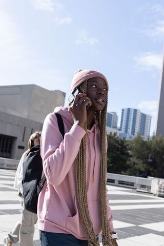 African american woman talking on phone wearing pink hoodie and backpack in urban plaza