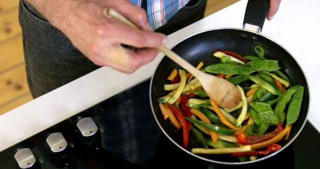 Person stir-frying fresh, colorful vegetables in a nonstick pan using a wooden spoon. Ideal for use in food blogs, healthy eating articles, cooking guides, recipes, culinary magazines, and advertisements for kitchen utensils or cookware.