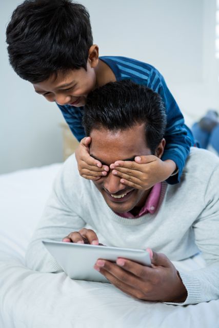 Father and son enjoying playful moment in bedroom. Son covers father's eyes while father uses tablet. Perfect for family, technology, and bonding themes. Ideal for parenting blogs, family-oriented advertisements, and technology usage in family settings.