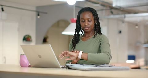 African woman checking smartwatch while working on laptop in modern coworking space