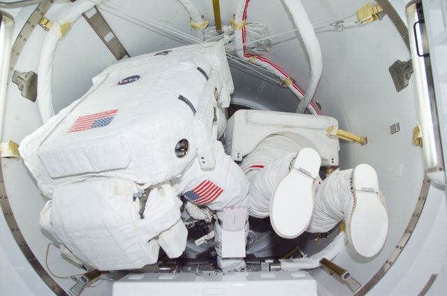 This image showcases astronauts preparing for an extravehicular activity (EVA) while in the Crew Lock compartment of their spacecraft. Both are dressed in full spacesuits, featuring the American flag. This image can be used to illustrate advanced space technologies, the human side of space missions, or educational material on space exploration and astronaut activities.