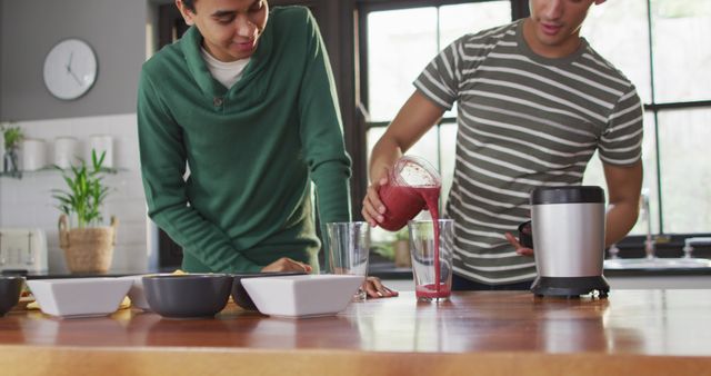 Two friends are in a kitchen making smoothies together. One friend is pouring the smoothie into a glass while the other is smiling. The image exudes a casual, healthy lifestyle vibe, ideal for use in themes related to healthy eating, cooking at home, friendship, and young people socializing in home settings.