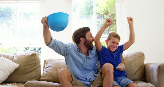 Father and son raising arms in excitement while sitting on couch in living room. Perfect for concepts related to family bonding, home entertainment, father-child relationships, and celebrating moments together.
