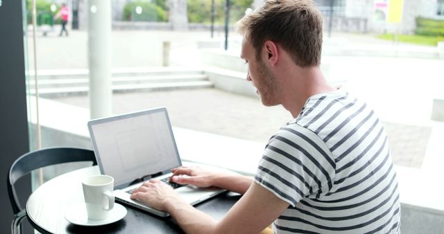 Young man sitting in cafe working on laptop. Casual setting with coffee on table. Ideal for illustrating freelance work, remote job concepts, and modern business trends.