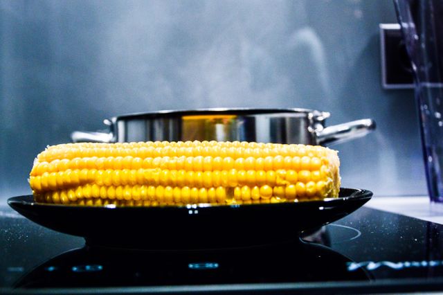 Vibrant corn on the cob on a sleek black plate, with a cooking pot visible in the background, creating a modern kitchen vibe. Ideal for culinary blogs, healthy eating advertisements, and kitchenware catalogues.