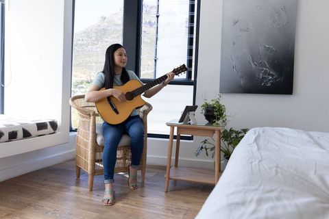 Asian woman playing acoustic guitar in minimalist serene sunlit bedroom near large window