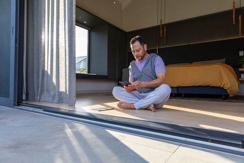 Mature asian man sitting by open patio using smartphone and tablet in sunlit bedroom