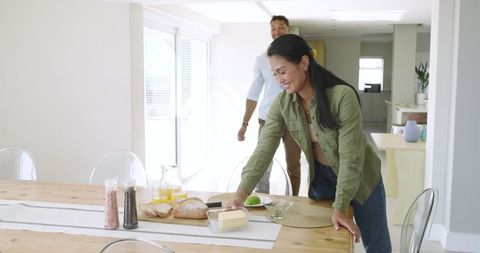 Couple Preparing Bright Modern Breakfast While Woman Places Butter on Wooden Table, Serving Juice