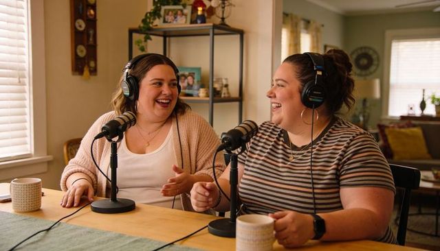 Two co-hosts recording cozy home podcast wearing headphones and chatting over microphones