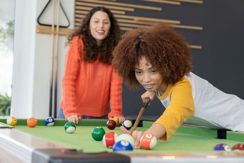 Young women playing pool in modern rec room enjoying friendly billiards, sunlit casual leisure momen