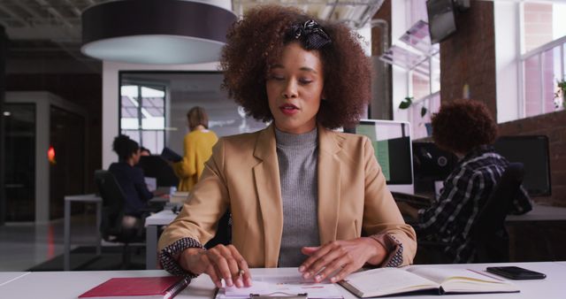 Confident businesswoman sitting at office desk, working on documents with focus. Team members collaborating in background in modern office setting. Ideal for business, teamwork, and corporate themes, representing professionalism, corporate culture, and workplace dynamics.