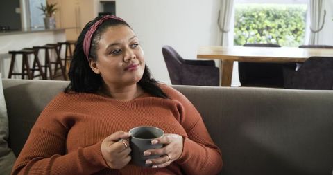 Contemplative woman relaxing on couch holding mug in cozy modern home interior