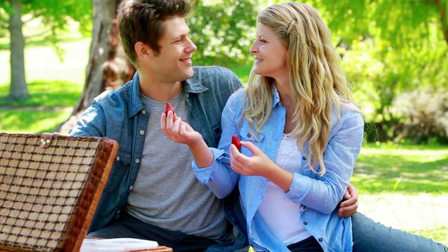 Couple smiles and enjoys strawberries while having a picnic in lush green park. Suitable for themes of love, romance, outdoor activities, summer relaxation, and healthy eating.