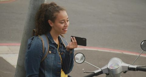 Young woman leaning on curb smiling and speaking into smartphone next to parked scooter