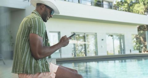 Relaxing poolside and texting on smartphone at luxury modern villa with sunglasses