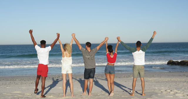 Group of friends from diverse backgrounds standing in a row holding hands and lifting their arms in celebration. Use for themes of friendship, diversity, unity, summer vacations, and beach leisure activities.