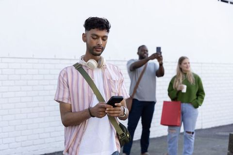 Young indian man using smartphone with headphones while friends socializing on urban sidewalk
