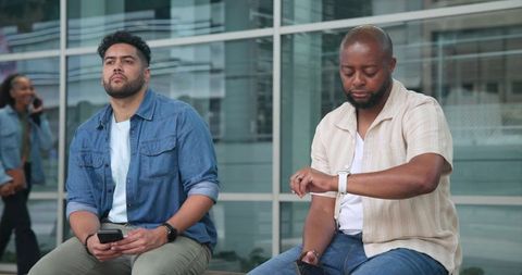 Diverse coworkers sitting outdoors checking smartwatch and smartphone at modern glass plaza