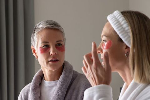 Mature women applying under-eye gel patches during cozy at-home skincare routine