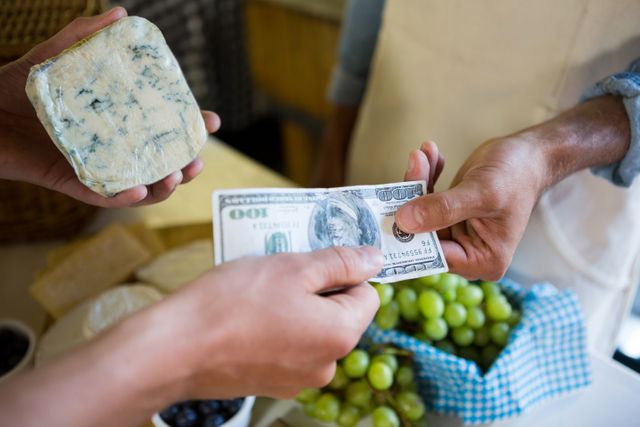 This image shows a customer paying with cash at a farmers market. The vendor is receiving the payment while holding a piece of cheese. Fresh grapes and other produce are visible in the background. This image can be used for topics related to local markets, organic food, retail transactions, and supporting small businesses.