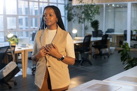 Confident professional woman standing, gazing in modern open-plan office wearing beige vest