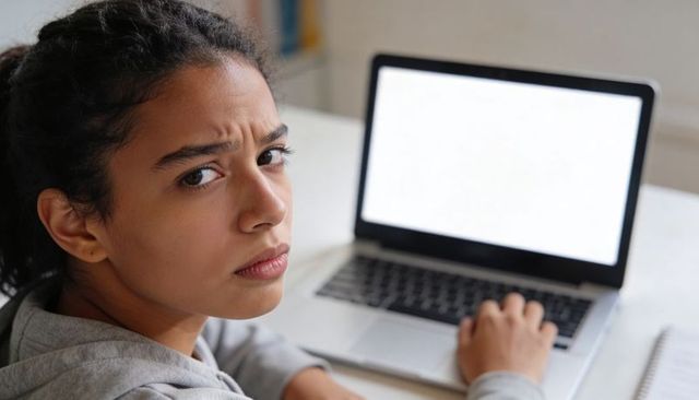 Frustrated Teen Girl Studying on Laptop at Home Desk with Blank Screen and Notebook