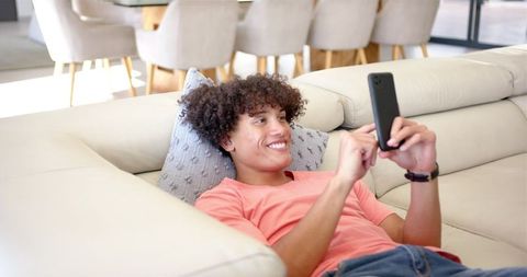 Smiling Young Man with Curly Hair Relaxing on Sofa Using Smartphone