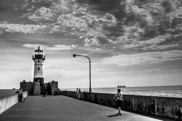 Calm sunset scene capturing a lone lighthouse with pedestrians walking and cycling along the pier. All captured in a black-and-white setting adding a nostalgic feel. Sky full of clouds adding texture to serene ocean view. This image is ideal for use in maritime-related projects, travel blogs, inspirational posters, or any design focused on solitude, peace, and nautical themes.