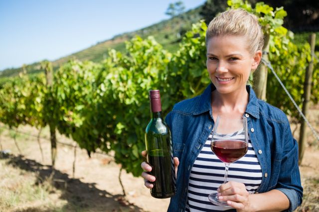 Smiling female vintner holding a wine bottle and glass in a vineyard on a sunny day. Ideal for use in articles or advertisements related to winemaking, viticulture, wine tasting events, rural tourism, and agricultural practices.