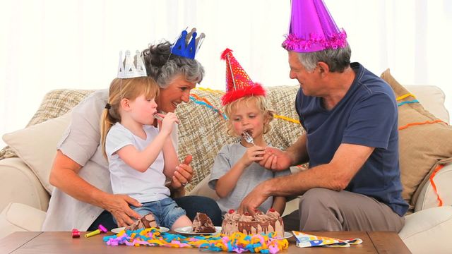 Grandparents and grandchildren are celebrating a birthday together at home. They are wearing party hats and eating cake, sharing joyful moments. Perfect for family-oriented brands, birthday party invitations, or articles promoting family relationships.