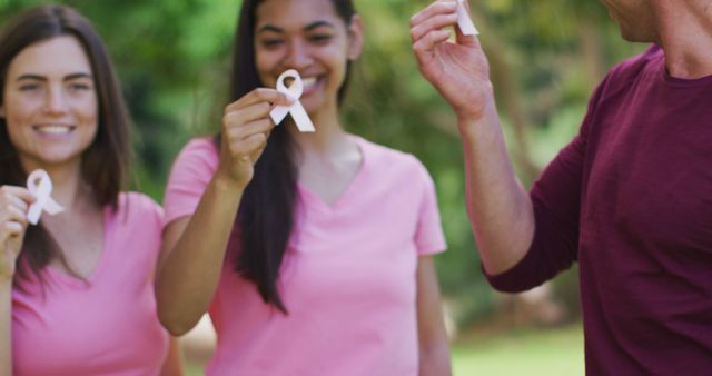 Portrait of diverse group of men and women smiling and holding breast cancer ribbons in park. health and positivity, breast cancer awareness campaign.
