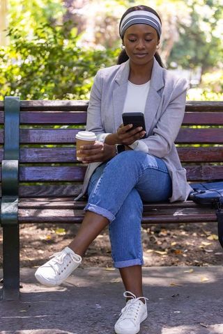 African woman holding coffee and scrolling smartphone on park bench in sunlit urban park