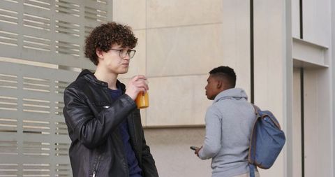 Two men standing by modern building sipping juice and checking phone on urban commute