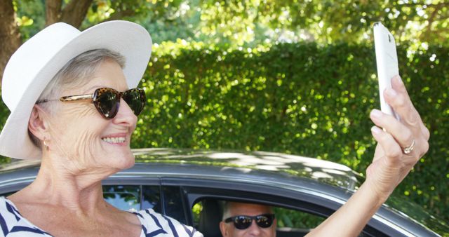 A senior Caucasian woman wearing a sunhat and sunglasses smiles as she takes a selfie, with a middle-aged man in the background sitting inside a car. Capturing joyful moments on a sunny day, the image reflects leisure and connectivity across generations.