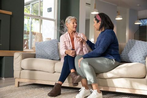 Older women chatting on sunlit living room sofa, cozy conversation and friendship moment