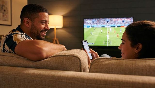 Couple watching live soccer on tv while checking smartphone and chatting on cozy beige couch